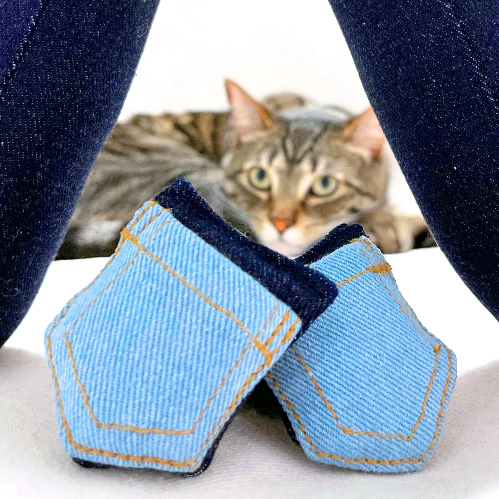 Tabby cat sitting and peaking out between two denim pocket cat toys on a light background.