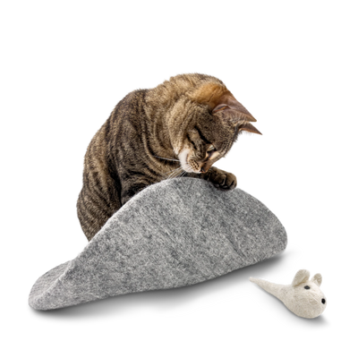 Tabby cat playing with a white felted wool mouse toy on a grey wool mat on a light background.