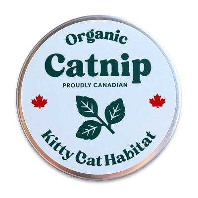 A round metal tin lid with a white label reading 'Organic Catnip' and 'Kitty Cat Habitat,' featuring green text and leaf graphics, red maple leaves, and 'Proudly Canadian' text, on a black background.