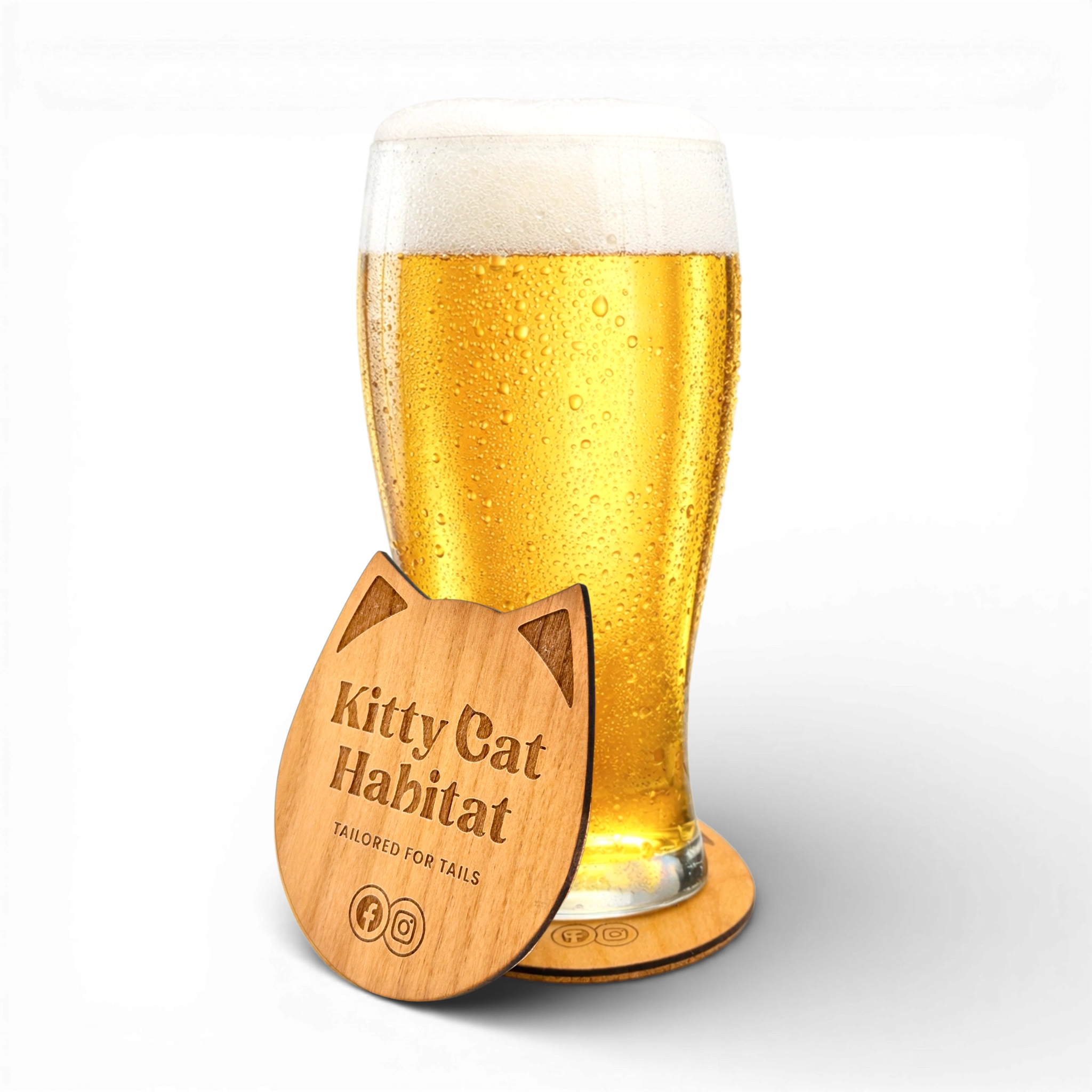 Beer glass resting on a wooden cat head drink coaster with a second Kitty Cat Habitat logo coaster in front on a light background.