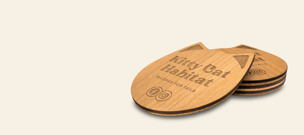 Stacked laser-engraved wooden cat-shaped coasters on a light cream background. The top coaster is fully visible, showing the text 'Kitty Cat Habitat' and 'TAILORED FOR TAILS', while a stack of coasters is partially visible underneath.