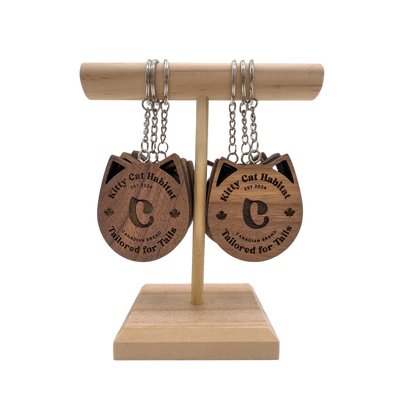 Several walnut wooden cat head keychains showing the logo side hanging on a wooden display stand on a light background.