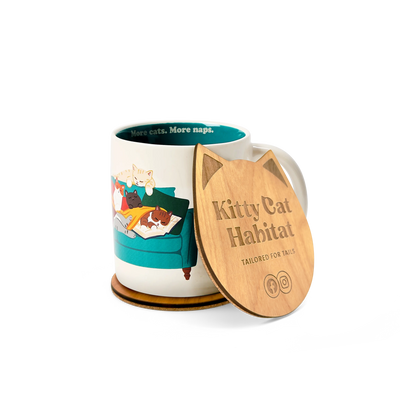 A ceramic mug with a colorful cat-themed illustration sits beside a wooden cat head shaped coaster. The lid is engraved with the text 'Kitty Cat Habitat' and 'TAILORED FOR TAILS'.